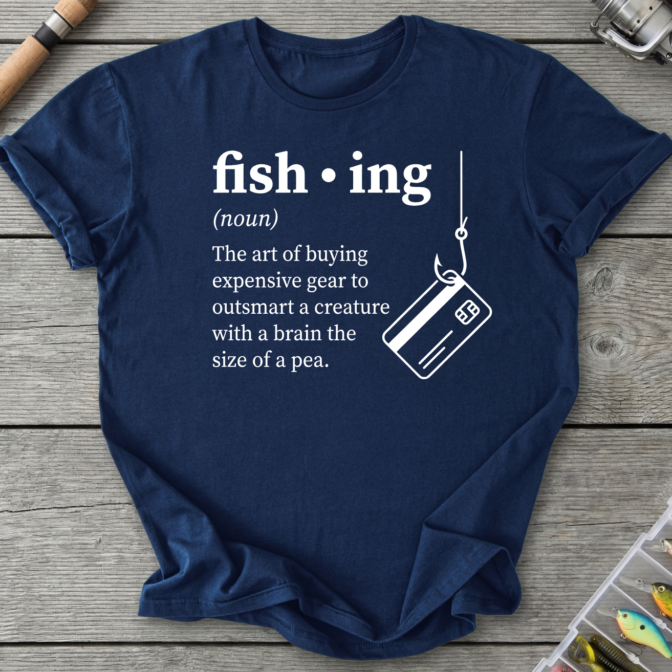 Navy blue t-shirt with fishing-themed text and graphic on a wooden surface with fishing equipment.